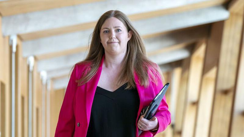 Gillian Mackay wears a pink blazer and black top and walks down a corridor in the Scottish parliament holding a laptop and notepad