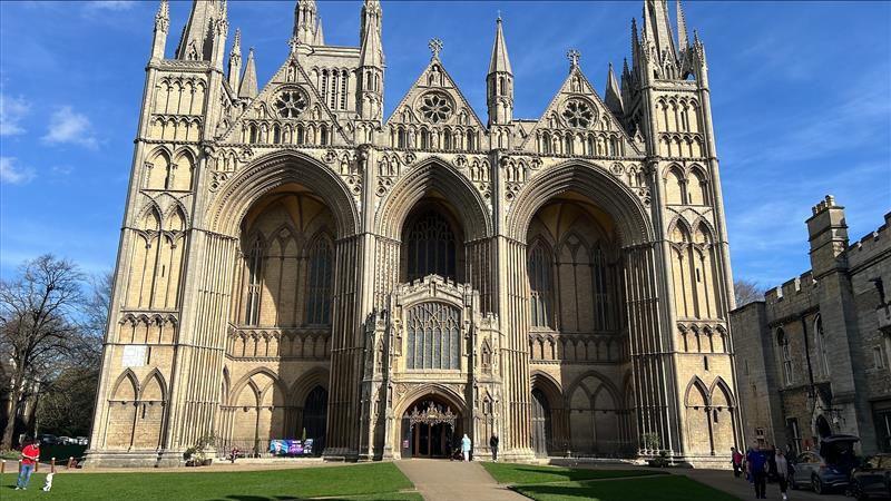 The outside of Peterborough Cathedral in Peterborough city centre. The cathedral is a light brown colour and sits on a patch of grass. The sky is blue.