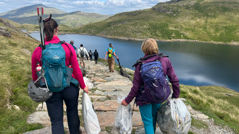 A group of volunteers, with two women in the forefront, walking along a path with their backs to the camera. Their is a grassy verge to the left, and a body of water to the right, and mountains in the distance. The volunteers are laden with full rubbish bags and litter pickers.
