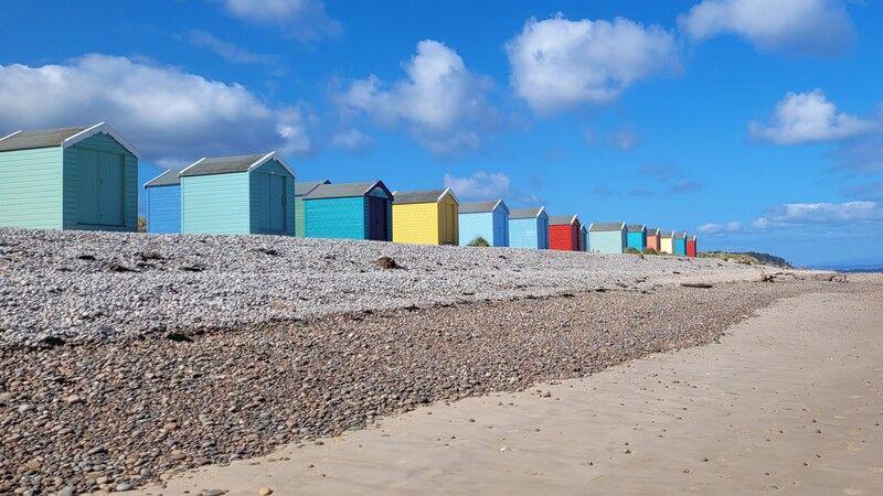 Colourful beach huts line up along a pebbly beach that becomes sandy closer to the water.