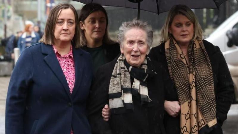 Four members of the family are standing underneath a large black umbrella. They have sad facial expressions and are all wearing coats and scarves.
