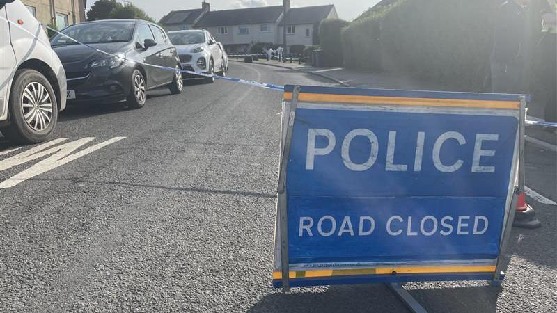 A blue sign which reads 'police road closed' placed on a residential street with a row of cars parked to the left hand side