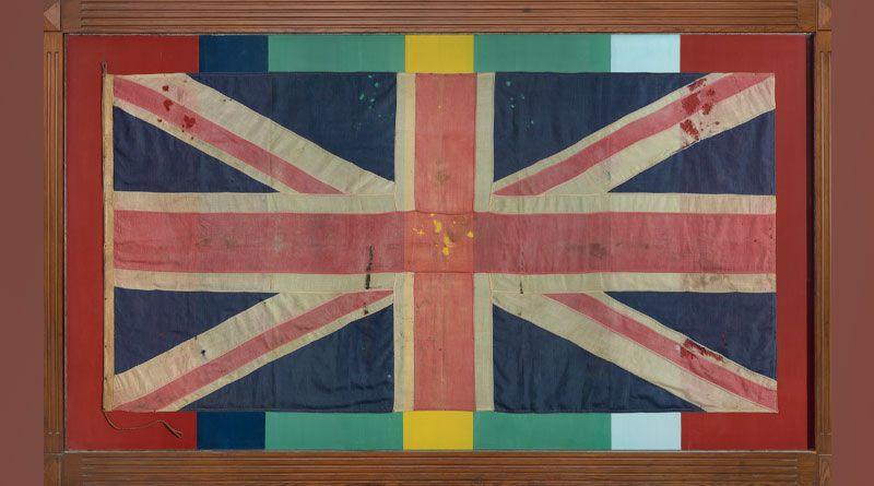 The Leiston FEPOW Memorial. It is a World War Two-era faded union jack which has been pinned to a background painted in the colours of the medal the Pacific Star: red, blue, green, yellow and white stripes and encased in a wooden frame. 