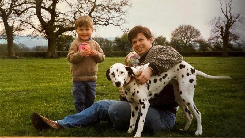 An archive picture of Dr Nicholas Bennett when his son Alexander was a boy. They are outside, in what looks like the countryside. Dr Bennet is sitting on the ground and playing with a Dalmatian. Alexander is holding a rugby ball and smiling for the camera.