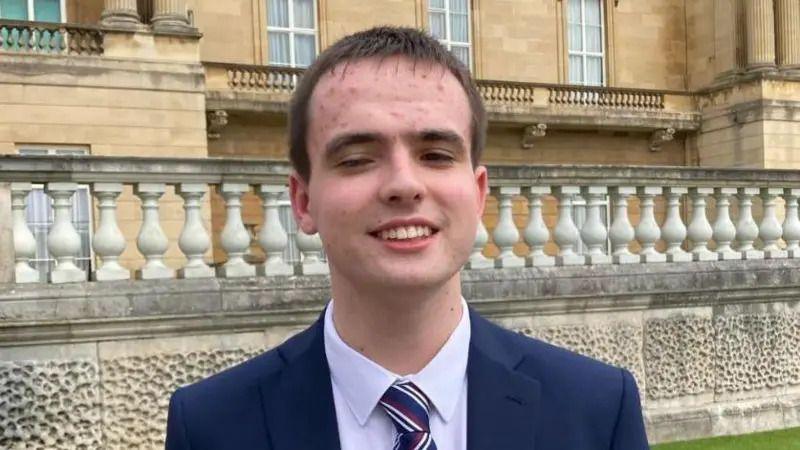 A young man wearing a navy jacket, white shirt and striped blue, white and red tie. He has short brown hair and is smiling. In the background is a grand looking building, with columns.