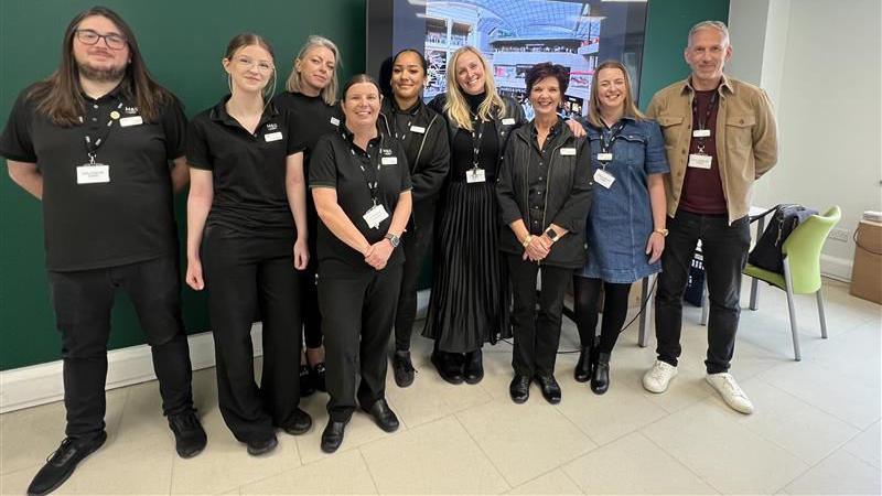 A group of eight people line up against a dark green wall inside the new M&S store in Bristol. They are mostly women but there are two men too. All of them apart from one man are in the store's dark uniform, and are wearing name tags