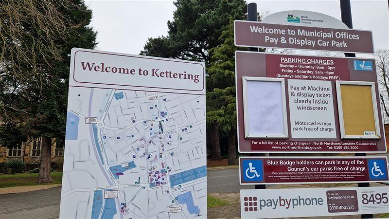 Two Kettering signs in a car park, including a welcome to Kettering map, and a details of a parking charges. There are trees and a road behind the signs.