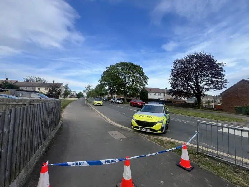 A police cordon blocks part of the pavement,. There is a police car blocking the road and other police car further away from the camera up the road. 