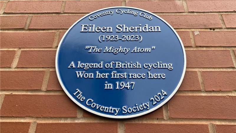 a blue circular plaque on a red brick wall that says " Eileen Sheridan (1923-2023) 'The mighty Atom' A legend of British cycling won her first race here in 1947". At the bottom of the plaque it reads "The Coventry society 2024"