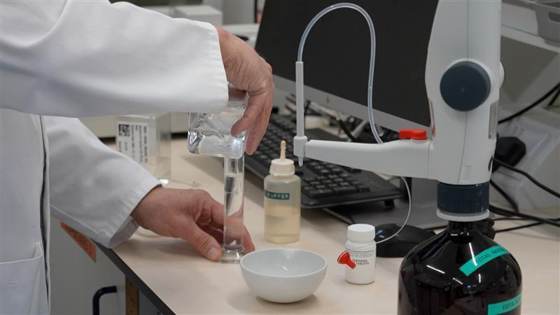 Someone in a lab testing water quality. The items are on a desk with a desktop behind it. The man doing the testing is wearing a white jacket.