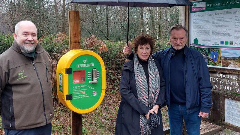 The defibrillator installed at the Ashdown Forest Centre before it was vandalised and stolen; one man is pictured to the left, while there is a woman and man pictured to the right. The man is holding an umbrella over both of them.