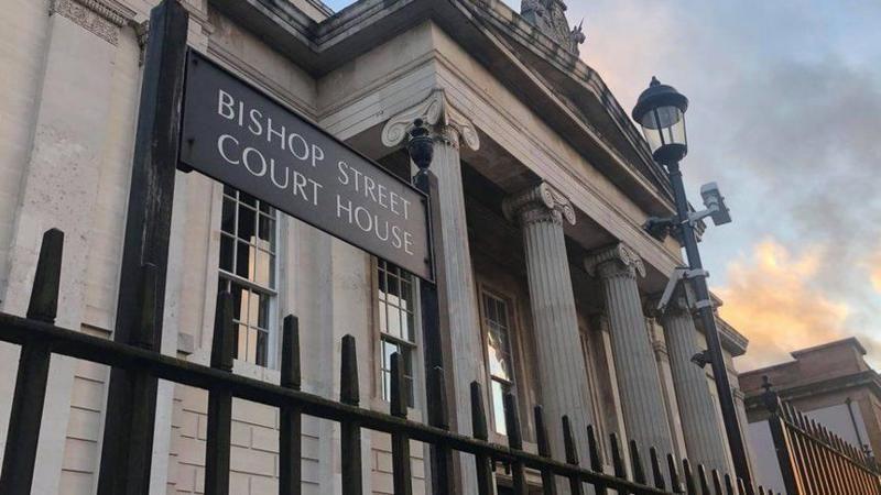 The front of the courthouse in Derry, A sign, saying bishop street courthouse can be seen through a line of iron railings, stands if front of the building's four large pillars..