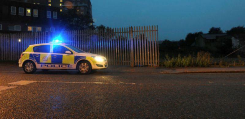 Police car parked at night. There is a silver metal fence behind it and a brick building in the background.