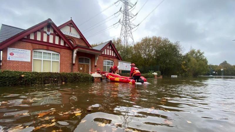 A church building with a red-beamed roof with water up to the front door and a red fire service boat with three firefighters by the door