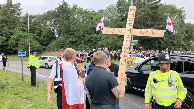 A man holding a large wooden cross that reads 'stop the boats now' with two St George flags attached on either sides of them. There are police officers also at the scene
