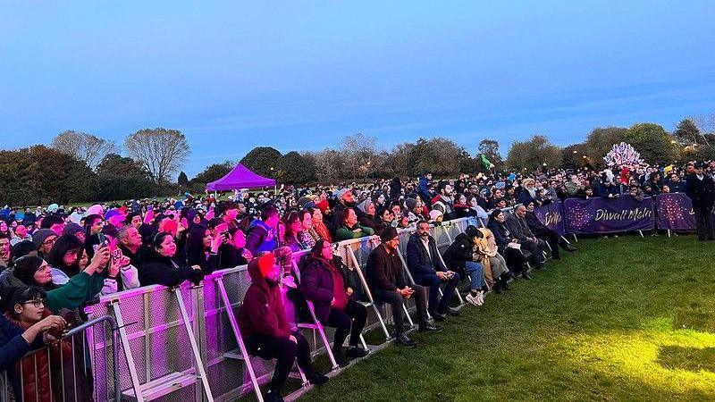 A crowd of hundreds of people gather at the Diwali Mela celebrations in Phoenix Park in 2023. Hundreds of people are standing behind a silver metal barrier, with around a dozen people sat in front of the barrier against a backdrop of a blue sky and grass.