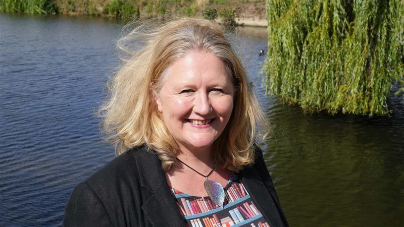A woman with thick, short blonde hair smiles and stands in front of a pond. She wears a black blazer with a top that has books on it.