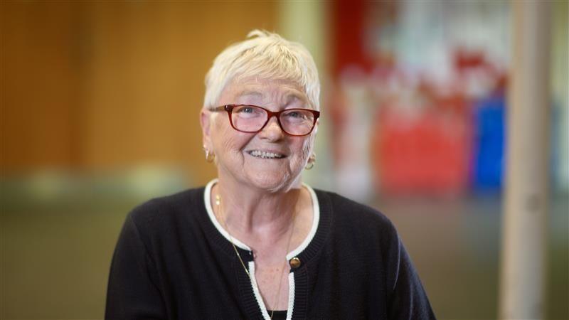 Ms Bird is smiling at the camera. She is wearing red glasses, a necklace and earrings. She has short white hair and is also wearing a blue and white cardigan. Her background is blurred.