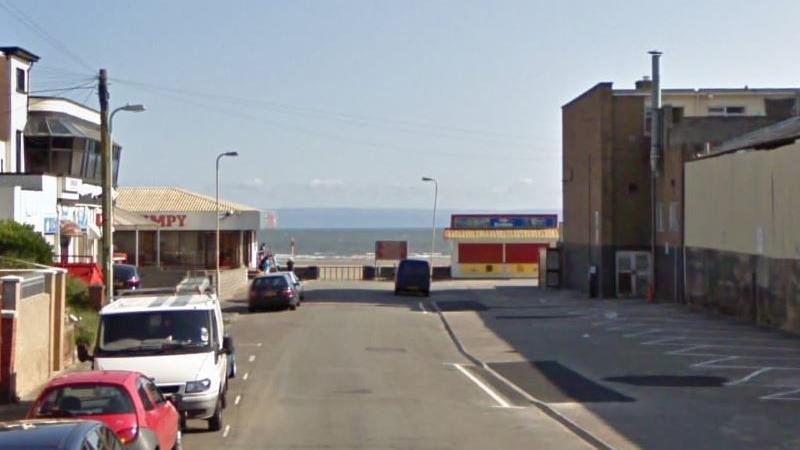 The end of Mackworth Road, leading to the beach and the sea at the end. Vehicles are parked on the left hand side, with empty parking spots to the right. A Wimpy restaurant can be seen in the background. 
