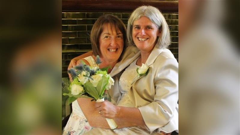 A woman with white hair holding flowers and sitting on the lap of another woman, who has brown hair.