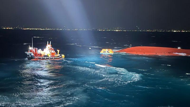 The orange upturned hull of a boat is visible in the water. It's nighttime and the waves are illuminated by lights. People are seen in a small lifeboat, plus another boat nearby.