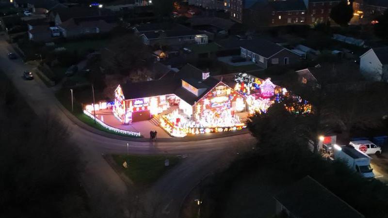 The couple's house is photographed at night-time from the air using a drone. It is covered with lights and decorations. It is a large detached house on a corner plot on a residential street.