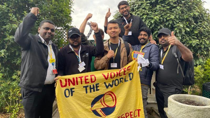Seven members of the cleaning and porters team at St Helier and Epsom Hospital hold up a yellow banner with the name of their union, United Voices of the World, printed on it, as it each member of the group raises their hands in salute.