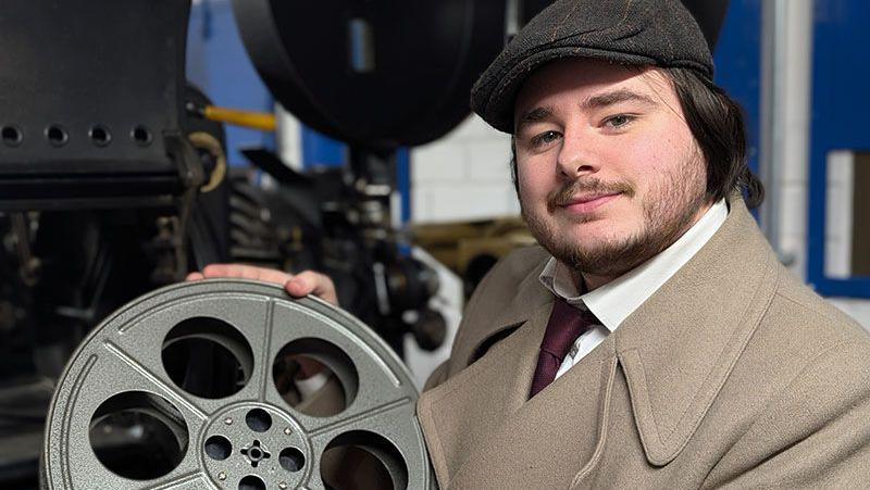 A man dressed in 1950s clothes, a smart coat with a shirt and tie and wearing a cloth cap, holds a film reel.