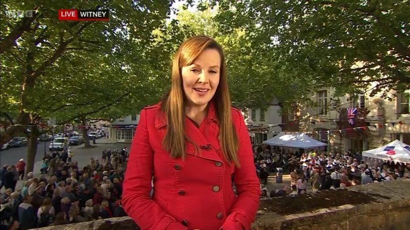 Alexis Green stands in front of crowds of people attending a street party to report on the weather. She has long light brown hair and wears a red trench coat.