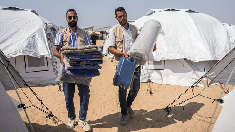 Two men carrying supplies including blankets and a water jug in a refugee camp in Gaza, surrounded by white circular tents on sand with a clear blue sky behind.