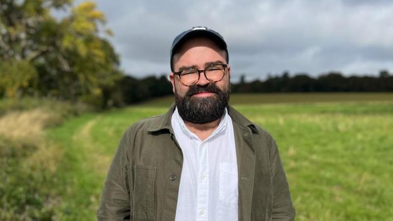 Jack Cornish standing in the corner of a tree-lined, grassy field. He is wearing a cap and glasses with a white shirt and dark green jacket.