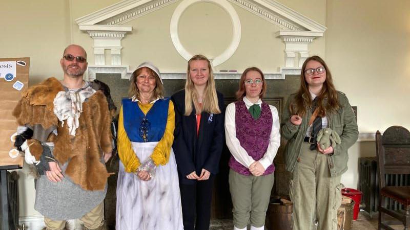 Members of the public dressed as characters from the series Ghosts flank a guide, pictured in front of a fireplace at West Horsley Place, the filming location for the series.