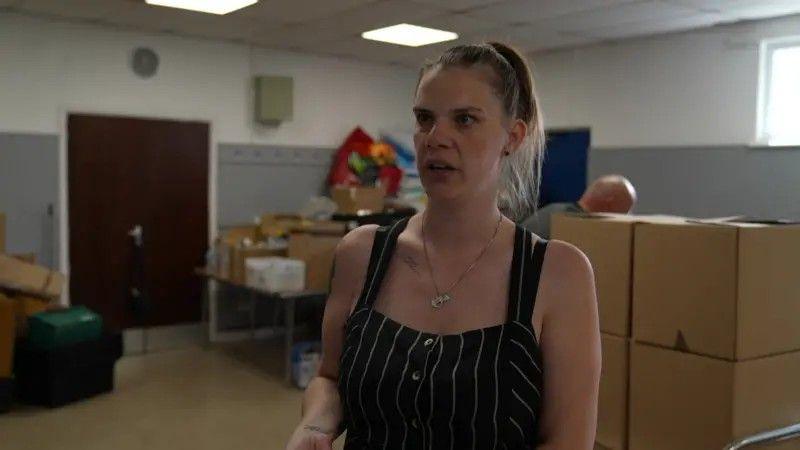 A woman with brown hair tied back and a black headband is wearing a black and white striped jumpsuit. There are brown boxes in a room behind her