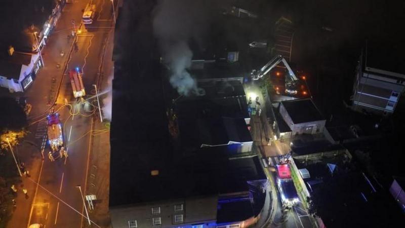 aerial shot of the scene of the fire, showing smoke from the building and fire engines on the street outside