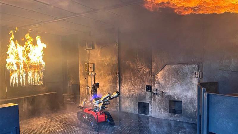 A red robot with road wheels and a silver hose approached a fire in a smoke-filled metal room. 