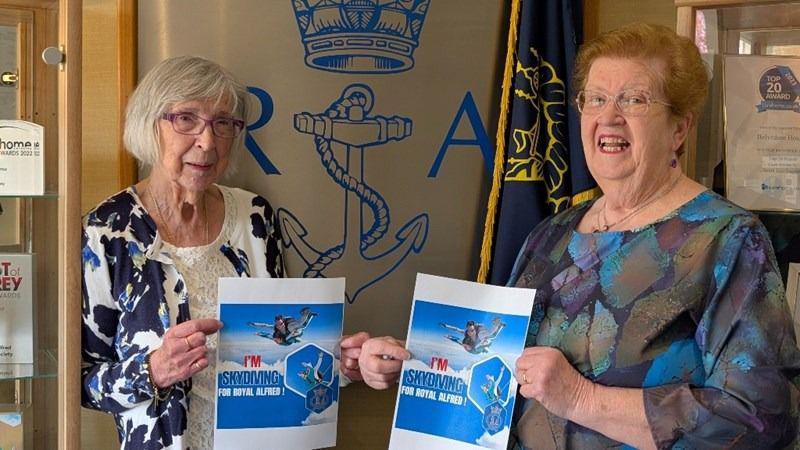 Joan and Liz stand holding a poster saying they are taking on a skydive.