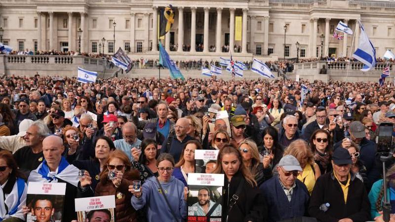A large crowd of men and women gathered in Trafalgar Square. Many hold or a draped in blue and white Israeli flags, and some Union Jacks. Others hold posters of some of the hostages, which features a picture of them, their ages and their names, printed in red, capital letters, in both English and Israeli. In the background more people can be seen lining the steps around the National Gallery.