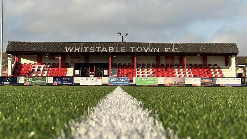 Whitstable Town FC's stand at its stadium has red seats and is seen from the level of the pitch.