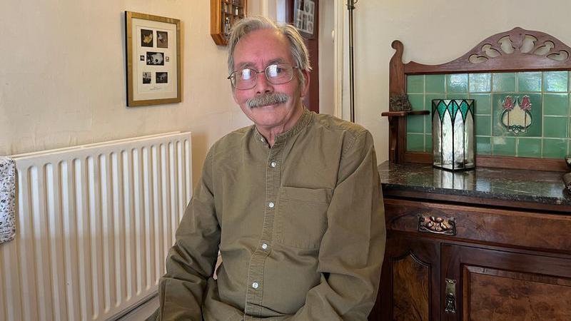 A man with short grey hair and a thick grey moustache with thin rimmed glasses is wearing a green long-sleeved shirt and sitting down looking at the camera. Behind him is a wooden vintage looking cabinet and to the left is a radiator and pictures on the walls.