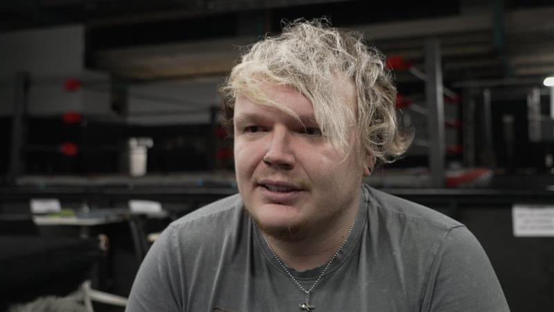 Lee Mitchell looks away from the camera while smiling. He has light hair that hangs over his forehead and wears a grey t-shirt with a chain around his neck that has a cross on it.