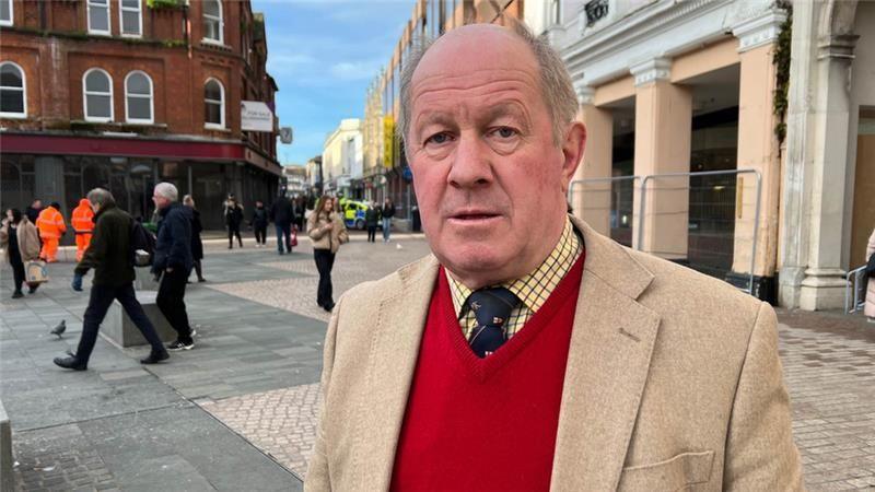 Tim Passmore is standing in front of buildings in Ipswich town centre. He has short grey hair and looks serious. He is wearing a yellow and black checkered shirt, a navy tie and a red V-neck jumper.