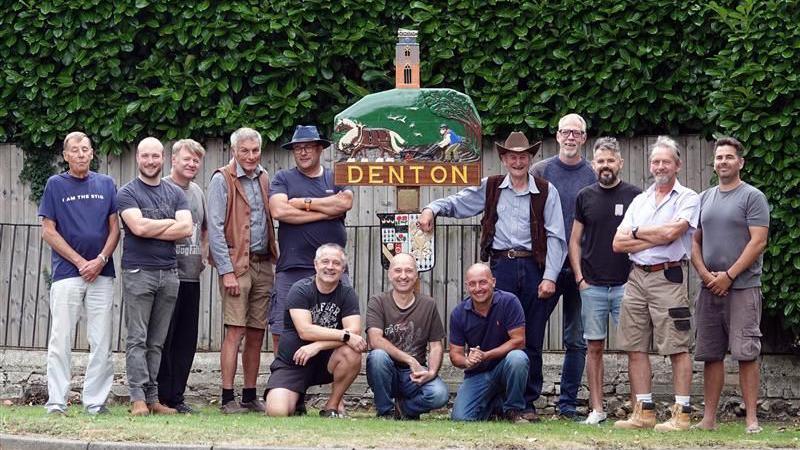 A group of men pose before a village sign. they are all smiling