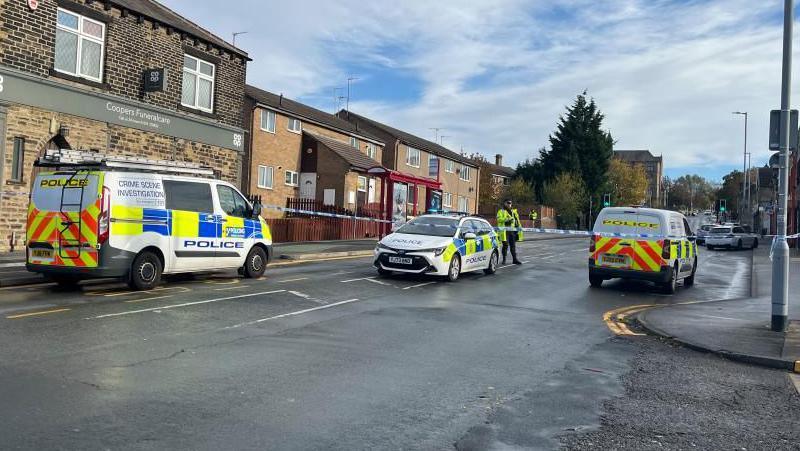 A police cordon in place on Barkerend Road in Bradford. A crime scene investigation van and two police cars are parked in the road, with police tape drawn across the street.