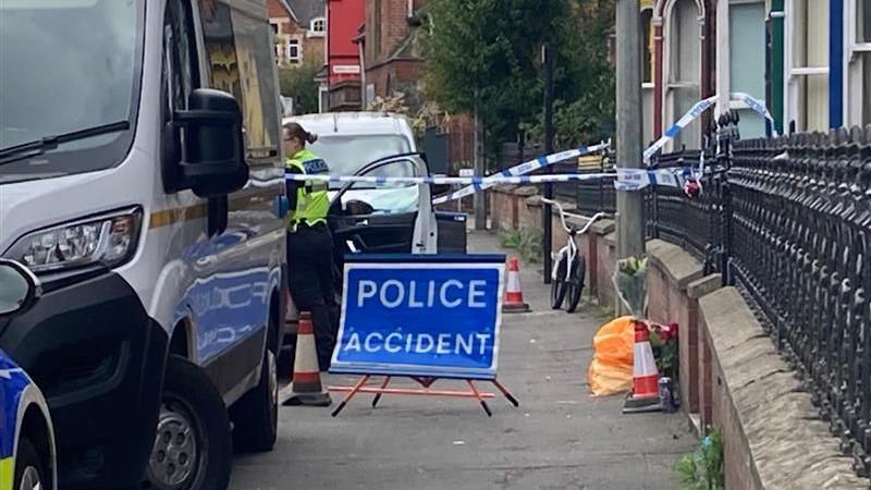 A blue police accident sign with blue and white cordons crossing a path. Vehicles are parked on the left. Houses are on the right of the path. A police officer can be seen behind the cordon.