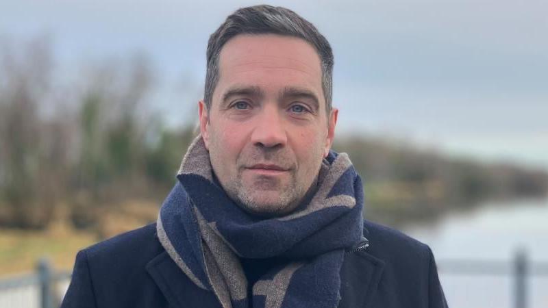 Michael Cadden looks into the camera wearing a large black and brown scarf and dark overcoat. He has black hair with grey coming through. He is standing in front of a body of water with guard railings around him.