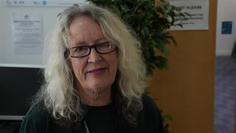 Carole Jones smiles at the camera. She has shoulder length grey hair and wears glasses and a dark top. A green plant sits behind her along with a white board with a piece of paper stuck on it with some information that is blurred.