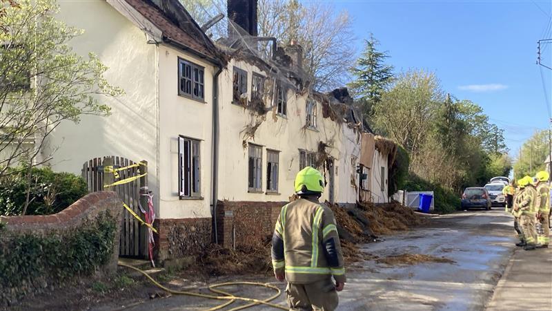On the left is a row of five adjoined houses that have thatched roofs. The fire has left the cream coloured houses with no roofs, and there is some fire damage to the front of the building. A fire officer is standing on the road outside of the houses.