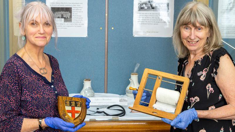 Two women standing in front of a display on a blue display board - the woman on the left is wearing a purple dotted top and a necklace and is holding a small shield with a St George's Cross on it. The woman on the right is wearing a black top with pink flowers and is holding a wooden ornament.