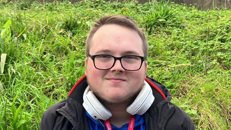 Brodie Graham stands in front of a grassy bank wearing dark glasses and a black coat. He has a pair of white headphones around his neck.