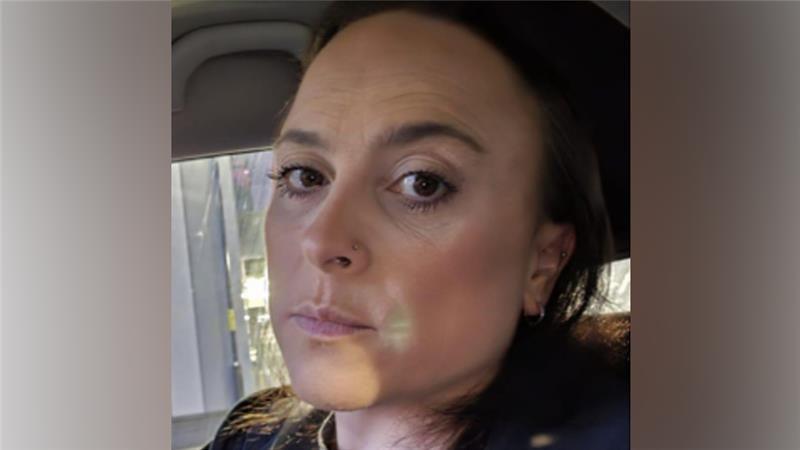 A woman with brown hair in a ponytail looking at the camera. She has a nose piercing and is wearing small silver hoop earrings. 